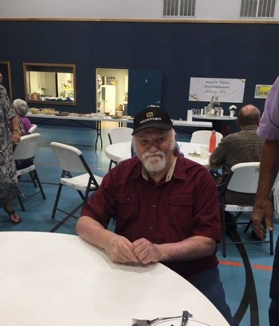 Frank, an older man with a gray beard and white hair, wearing a red short-sleeve shirt and a dark cap, sits at a round table in a bright indoor space
